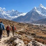 Three trekkers with backpacks walking along a rocky Himalayan trail with snow-covered mountains and stone buildings.