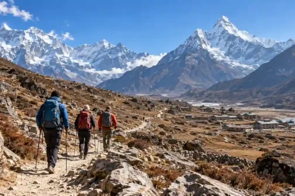 Three trekkers with backpacks walking along a rocky Himalayan trail with snow-covered mountains and stone buildings.